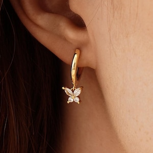 May include: Gold-colored hoop earring with a small butterfly charm. The butterfly is adorned with clear, sparkling stones. The earring is shown on a person's ear, with the focus on the jewelry's design and details.
