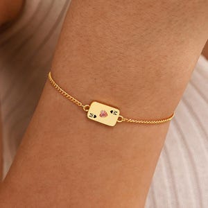 May include: A gold-colored bracelet with a rectangular charm featuring a heart and two small black shapes. The bracelet is on a delicate chain and is worn on a wrist.