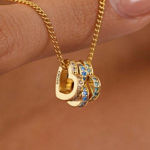 Puede incluir: Un collar dorado con tres colgantes en forma de corazón. Los colgantes están adornados con piedras preciosas azules y transparentes. El collar está sostenido por un dedo, destacando la fina cadena y el diseño del colgante. La palabra "Olivia" está grabada en uno de los corazones.
