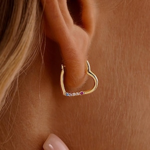 May include: A gold heart-shaped earring with small, colourful gemstones. The earring is worn on an ear, with a hand and hair visible in the frame. The gemstones are blue and red.