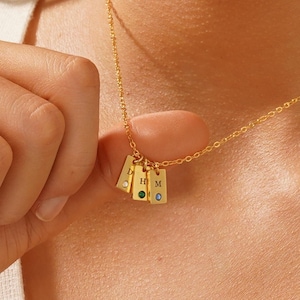May include: Gold necklace with four small rectangular charms, each with a letter engraved on it. The charms have small gemstones in different colors.