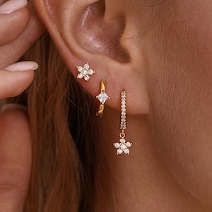 Op de afbeelding: Goudkleurige oorbellen met een bloemthema. De set bevat een kleine oorbel, een oorring met een vierkante edelsteen en een bungelende oorbel met een bloemhanger. De oorbellen zijn versierd met kleine, sprankelende kristallen.