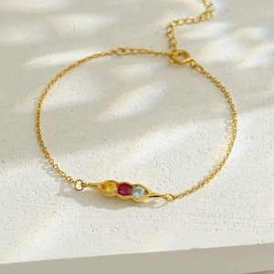 May include: A gold chain bracelet with a pea pod charm featuring three colorful gemstones. The charm is in the center of the bracelet, with the chain extending to either side.