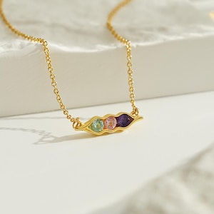 May include: A gold necklace with a pea pod charm featuring three colourful gemstones. The charm is in the shape of a pea pod with three round gemstones inside. The gemstones are green, pink, and purple.