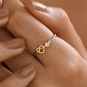 May include: A gold ring featuring a delicate heart-shaped knot design on one side and two small, circular bezel-set gemstones on the other. One gemstone is light pink and the other is light blue, both set into a thin, polished gold band.