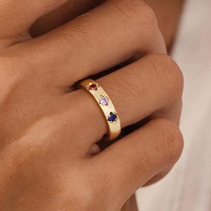 May include: A gold ring with three colorful gemstones: red, purple, and blue. The gemstones are set in a row along the ring's band. The ring is a simple, elegant design.