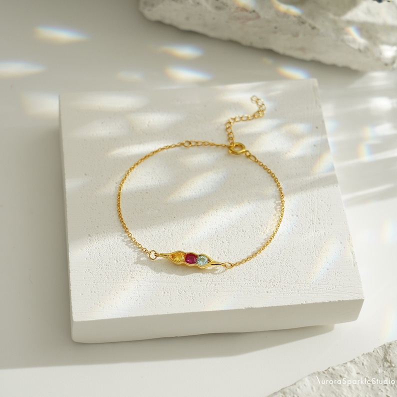 May include: A gold chain bracelet with a pea pod charm featuring three colorful gemstones. The charm is in the center of the bracelet, with the chain extending to either side.