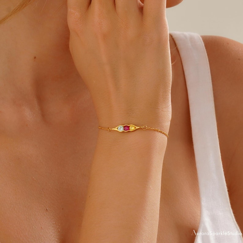 May include: A gold bracelet with a pea pod charm featuring three round gemstones: a light blue, a red, and a yellow.