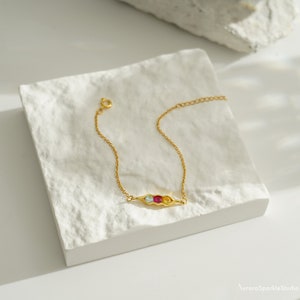 May include: A gold chain bracelet with a pea pod charm featuring three colorful gemstones: blue, red, and yellow.