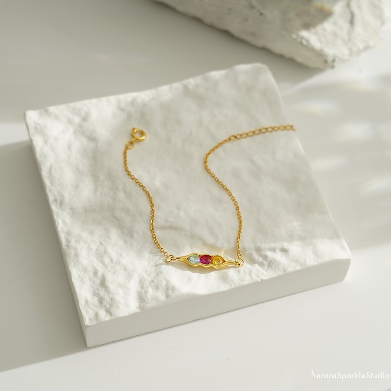 May include: A gold chain bracelet with a pea pod charm featuring three colorful gemstones: blue, red, and yellow.