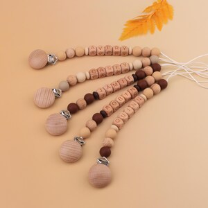 May include: Five wooden baby pacifier clips with names spelled out in wooden letter beads. The clips have a wooden disc and a metal clip. The beads are in shades of brown and beige. The names are Avery, Casey, Jamie, Robin, and Quinn.