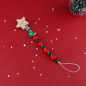 Peut inclure: Attache-tétine en bois en forme d'étoile avec le nom "Finn" gravé. Le clip comporte un charm en forme de sapin de Noël vert et est enfilé avec des perles rouges et vertes. Sur fond rouge avec des points blancs.