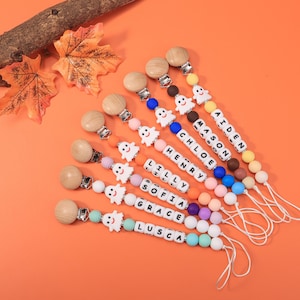May include: A collection of personalized baby pacifier clips with wooden beads, colorful silicone beads, and white ghost-shaped charms. Each clip features a name spelled out in white letter beads. The clips are arranged on an orange surface.