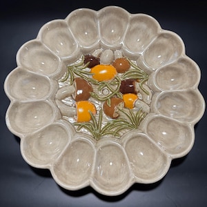 May include: A beige ceramic deviled egg serving platter with a scalloped edge. The center features a raised design of colorful mushrooms and green foliage. The platter has 18 egg-shaped indentations around the edge.