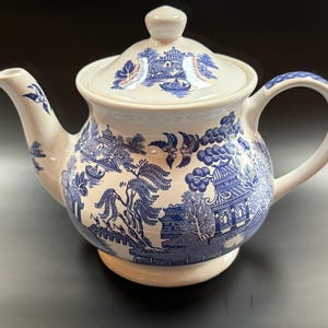May include: Cream-colored ceramic teapot with a blue Willow pattern design. The teapot has a rounded body, a curved spout, and a handle. The lid features a matching design and a small knob. A classic piece for tea service.