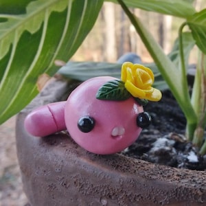 May include: A pink clay figurine of a worm with a yellow flower on its head. The worm has black eyes and a small mouth. The figurine is sitting on a brown pot.