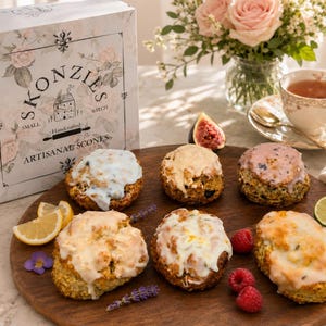 May include: A wooden serving board displays six artisan scones with various toppings, including white and pink icing. A box labeled "Skonzies" sits nearby, along with a cup of tea, flowers, and fruit. The scones are the focal point of this food-themed image.