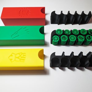 Könnte beinhalten: Drei farbige Boxen mit verschiedenen Symbolen auf der Vorderseite und zwei schwarze Organizer mit Fächern für kleine Token. Die Token sind grün und haben verschiedene Symbole.