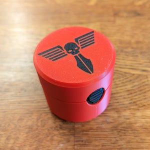May include: A cylindrical, red object with a black graphic on top featuring a skull with wings and a spear. The object has a small, circular opening on the side. The item is likely a container or grinder.