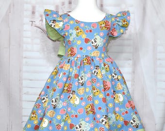 Può includere: Un vestito azzurro chiaro con un bordo verde e una stampa di orsi, fragole e mele. Il vestito ha maniche corte con volant e una gonna a pieghe.