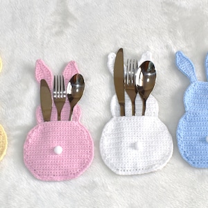 Puede incluir: Cuatro porta cubiertos de ganchillo en forma de conejo en amarillo, rosa, blanco y azul. Cada porta cubiertos tiene un pompón blanco en la parte inferior y contiene un tenedor, un cuchillo y una cuchara.