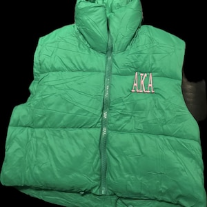 Puede incluir: Un chaleco acolchado verde con cuello alto y cremallera completa. El chaleco presenta las letras bordadas "AKA" en rosa en el pecho. El chaleco es de color verde liso.
