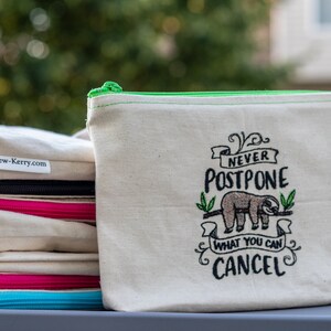 May include: A stack of canvas zipper pouches in natural tones. One pouch has a green zip and a sloth graphic with the text "Never Postpone What You Can Cancel". Other pouches have black, pink, and turquoise zips.