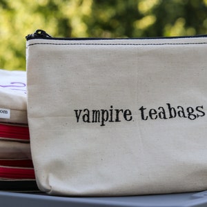 May include: A white canvas zippered pouch with black stitching and a purple embroidered design that says "vampire teabags".