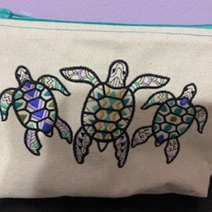 May include: A beige canvas zippered pouch with a teal zipper pull. The pouch features an embroidered design of three colorful sea turtles with geometric patterns.