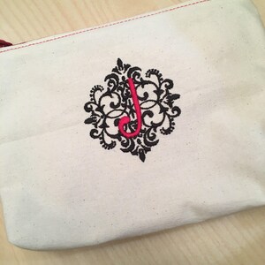 Puede incluir: Una bolsa de lona blanca con cremallera roja y un diseño bordado en negro con una letra roja "J" en un remolino decorativo.