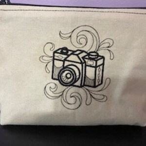 May include: A beige canvas zippered pouch with a black embroidered design of a vintage camera with swirling floral accents.