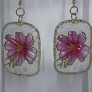 May include: A pair of gold earrings with a white background and a pink flower design.