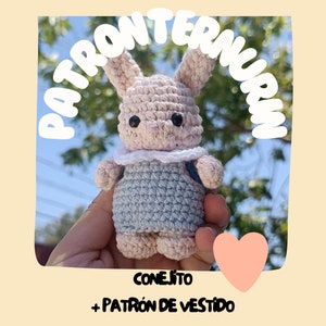 Puede incluir: Un conejito de crochet rosa con un vestido de crochet azul y blanco. El conejito está siendo sostenido en una mano. El texto "CONEJITO + PATRÓN DE VESTIDO" está debajo del conejito.