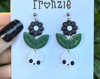 Puede incluir: Pendientes colgantes con una flor negra de centro blanco, una hoja verde con detalles grabados y una calavera blanca con cuencas oculares negras. Presentados en una tarjeta blanca con la inscripción Fronzie Polymer clay Studio.
