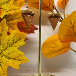 Può includere: Un paio di orecchini pendenti con una fetta di torta di zucca in miniatura. Gli orecchini sono realizzati in pasta polimerica e hanno un aspetto realistico. Gli orecchini sono esposti su un supporto dorato su uno sfondo di foglie autunnali.