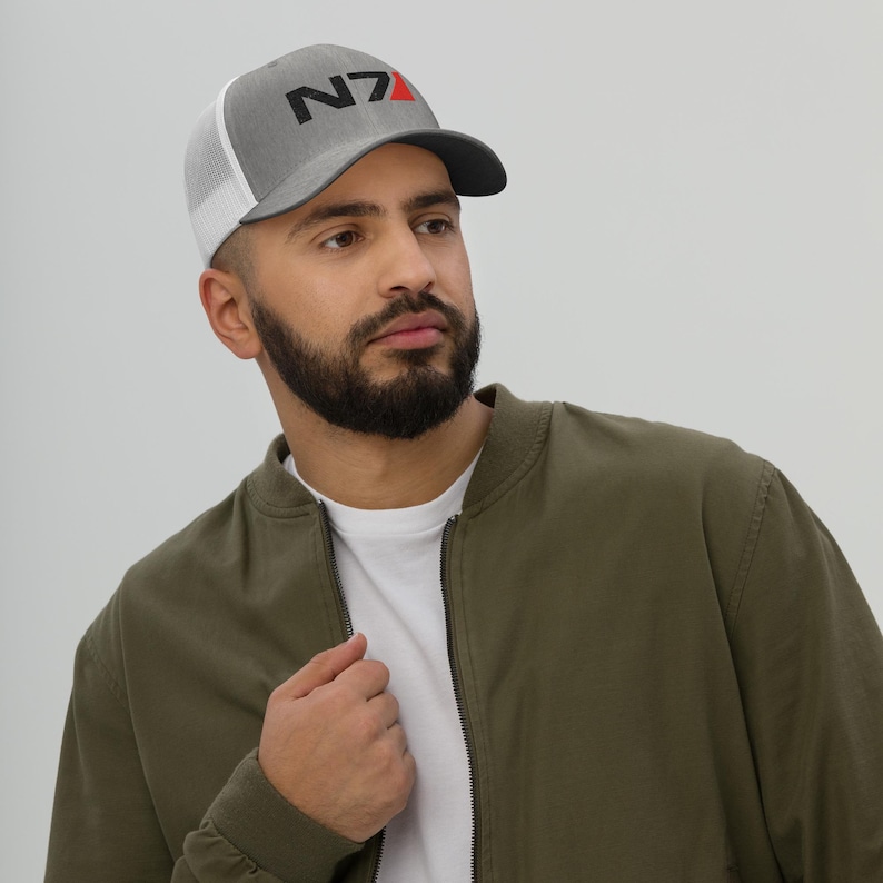 May include: A gray and white trucker hat with a black and red N7 logo. The hat is worn by a person with a beard and a green jacket.