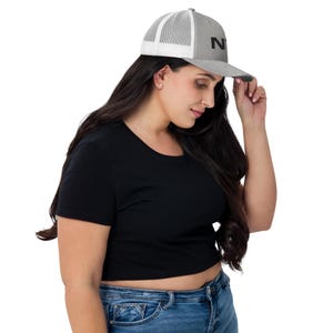 May include: A gray and white trucker hat with a black "NY" logo. The hat is worn by a person with long dark hair and a black shirt.