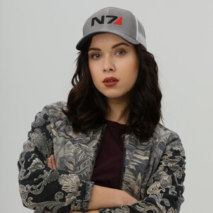 May include: A woman wearing a gray and white baseball cap with a black and red N7 logo. She is wearing a black and gray patterned jacket.