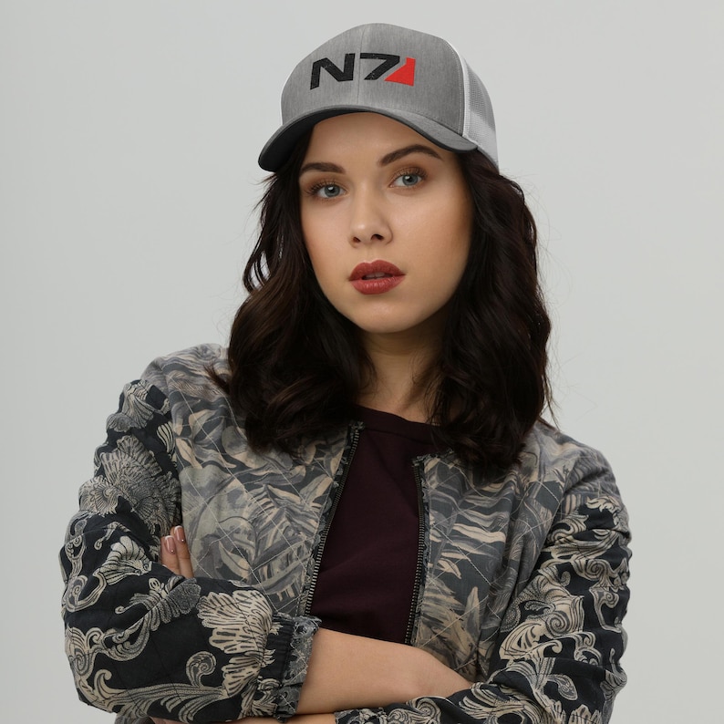 May include: A woman wearing a gray and white baseball cap with a black and red N7 logo. She is wearing a black and gray patterned jacket.