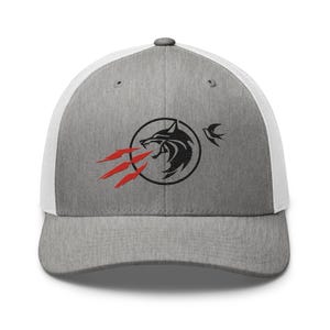 May include: Grey and white trucker cap featuring a black wolf logo within a circle, with red flame accents and a small bird. The cap has a curved peak and a mesh back.