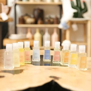 May include: A collection of small, clear glass bottles with white caps, each containing a different colored liquid. The bottles are arranged on a wooden surface, with labels visible. The background includes shelves with various items.