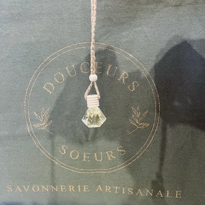 May include: A small, clear glass bottle with a gold lid hangs from a beige rope. The bottle is attached to a wooden bead. The bottle is hanging from a green canvas bag with the words "Douceurs Soeurs Savonnerie Artisanale" printed in gold.