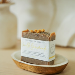 May include: A bar of brown soap with dried flowers on top. The soap is labeled "Douceurs de Soeurs" and "Vanille Gourmande". The soap is on a white oval dish with a brown wooden rim.