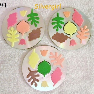 Puede incluir: Tres CDs con diferentes formas de fieltro de colores. Las formas son hojas, flores y otras formas abstractas. Los CDs están etiquetados con #1, #2 y #3. El texto "Silvergirl" está escrito en la parte superior de la imagen.