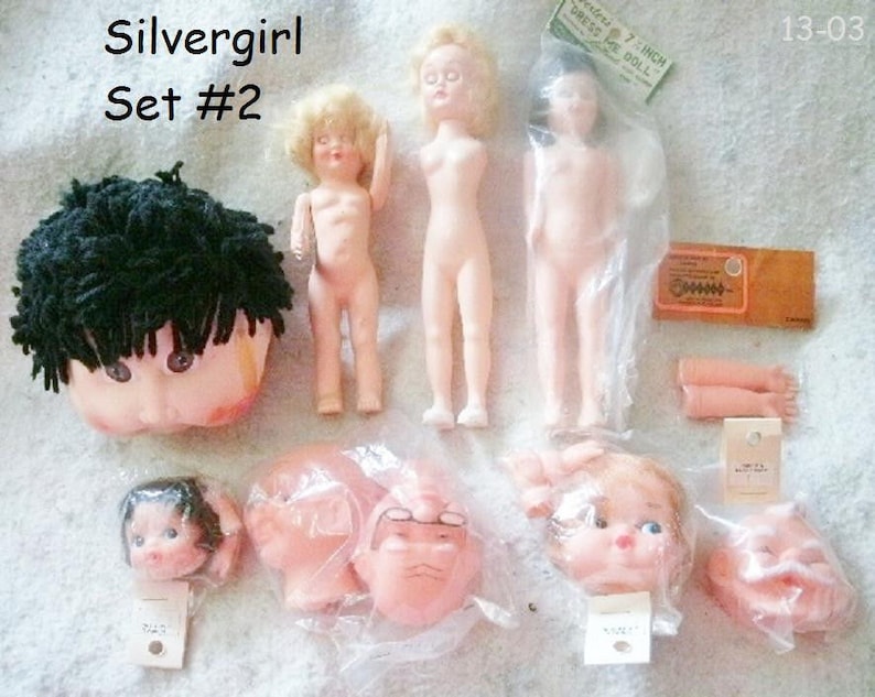 May include: A set of doll parts for a Silvergirl doll. The set includes a head with black yarn hair, a torso, two arms, and four different heads. The set is labelled "Silvergirl Set #2".