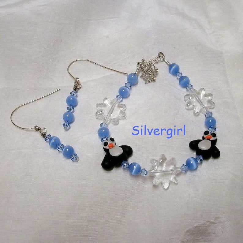 May include: A matching jewelry set featuring a bracelet and earrings. The bracelet and earrings have light blue beads, clear snowflake charms, and black and white penguin charms. The text "Silvergirl" is visible.