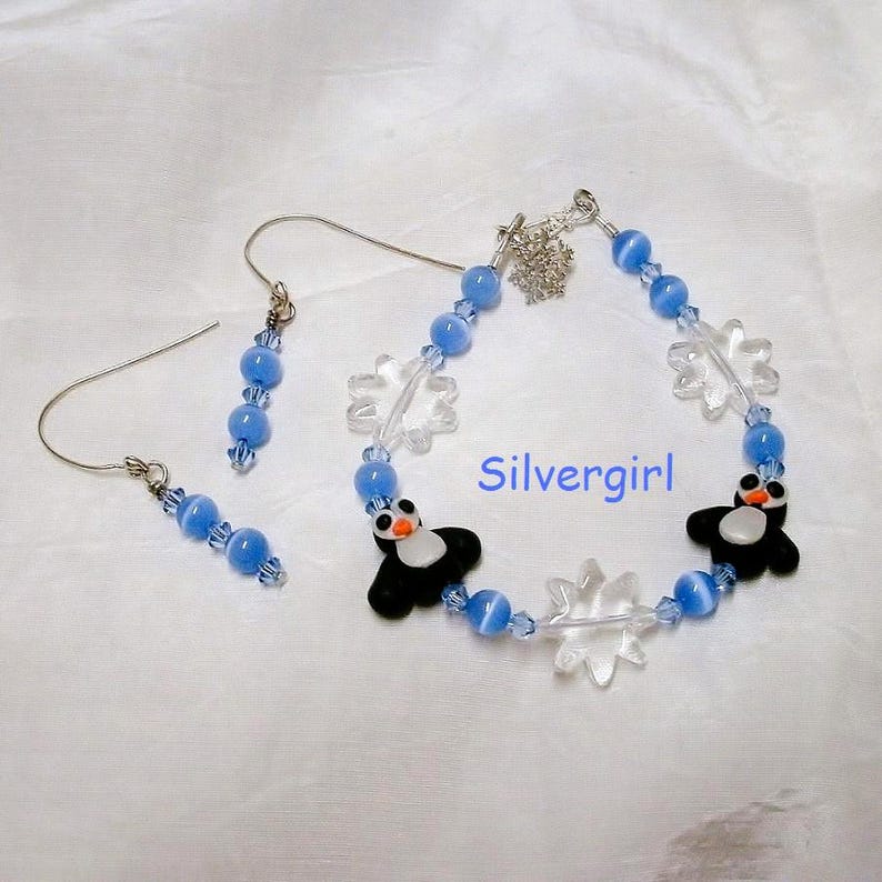 May include: A jewelry set featuring a bracelet and earrings. The bracelet and earrings have blue beads, clear snowflake charms, and black and white penguin charms. The text "Silvergirl" is visible on the bracelet.