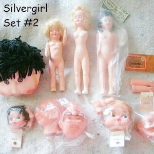 May include: A set of doll parts for a Silvergirl doll. The set includes a head with black yarn hair, a torso, two arms, and four different heads. The set is labelled "Silvergirl Set #2".