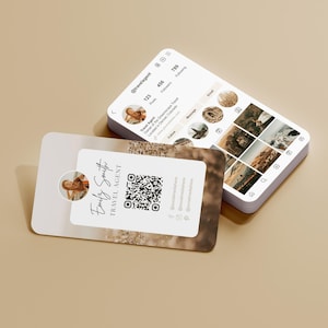 May include: A stack of white business cards and a smartphone displaying a travel agent's Instagram profile. The business card features a photo, name, and QR code. The phone shows travel photos and profile details.