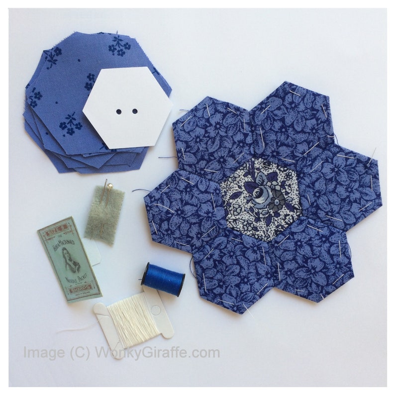 May include: A collection of quilting supplies, including blue floral fabric hexagons, a white paper hexagon, pins, thread, and a spool of white thread. The fabric features a blue floral pattern. The image suggests a crafting or sewing project.